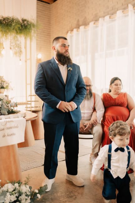 A young man with a full beard and short hair, dressed in a tailored navy blue suit with a white shirt and no tie, stands indoors during a celebration or event, with natural light streaming through she