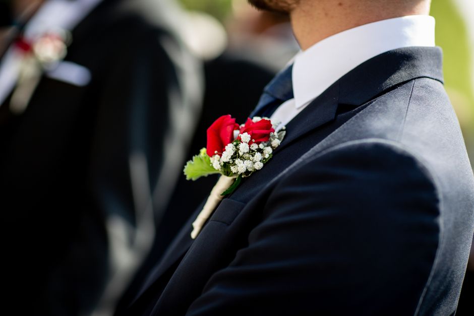 Close-up of a groom wearing a dark navy suit with a white shirt, adorned with a boutonniere featuring a single red rose, white baby's breath, and green foliage, pinned to the left lapel. The boutonnie