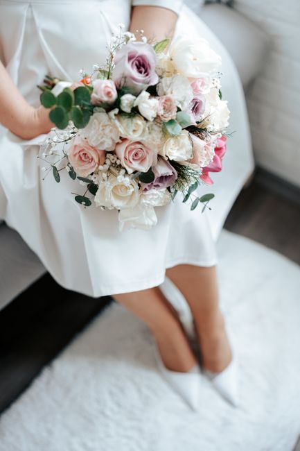 A person dressed in a white outfit is seated on a soft surface, holding a large, delicate floral bouquet close to their lap. The bouquet features an elegant arrangement of pale pink roses, white peoni