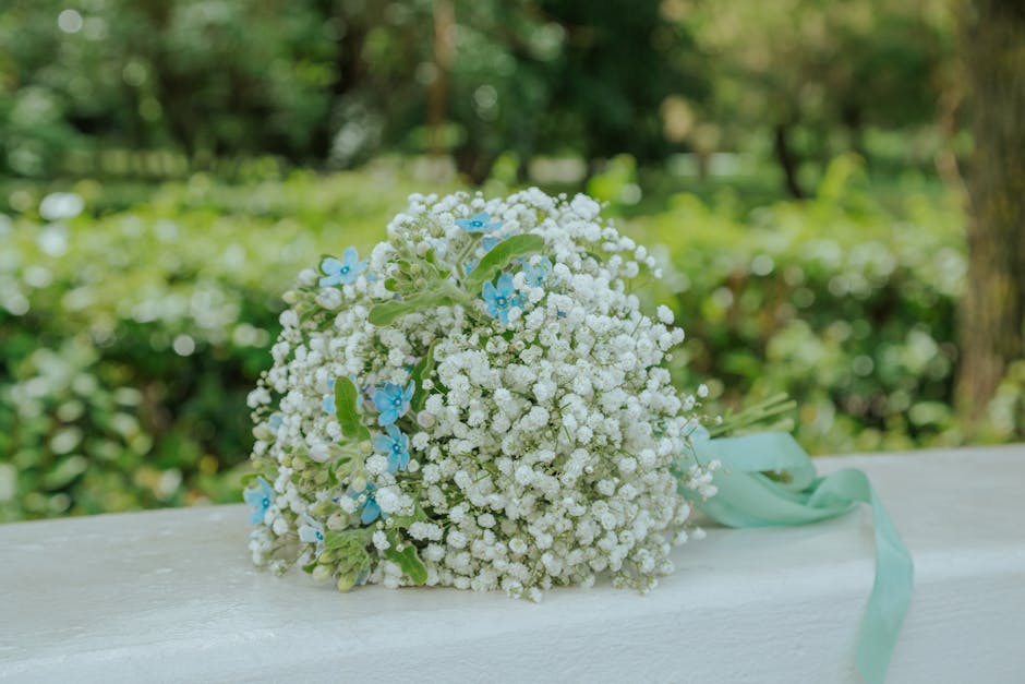 A bouquet of fresh, delicate white baby's breath (Gypsophila) flowers interwoven with small, light blue blossoms, arranged in a rounded floral arrangement with green foliage accents. The bouquet is ti
